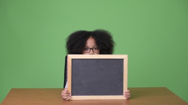 Jovem menina africana bonito com cabelo afro mostrando quadro negro enquanto sentado — Vídeo de Stock