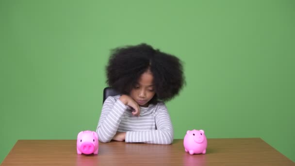 Young cute African girl with Afro hair choosing between two piggy banks — Stock Video