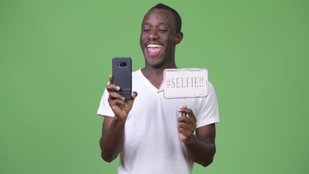 Молодой африканский мужчина делает селфи с бумажным знаком на зеленом фоне — стоковое видео