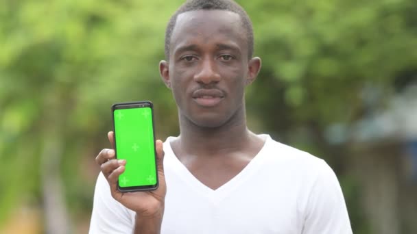 Jovem homem africano feliz sorrindo enquanto mostra telefone nas ruas ao ar livre — Vídeo de Stock