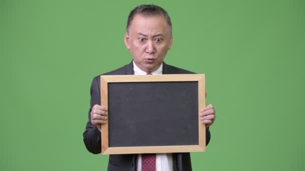 Mature Japanese businessman holding blackboard against green background — Stock Video