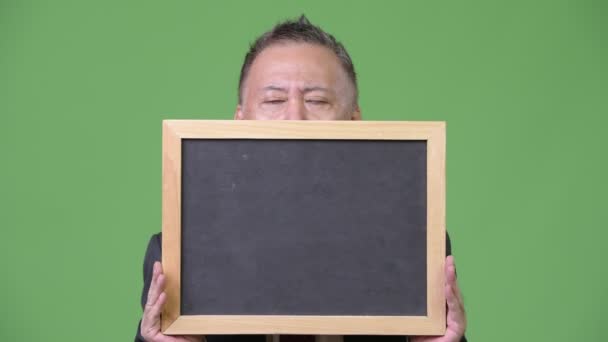 Mature Japanese businessman holding blackboard against green background — Stock Video