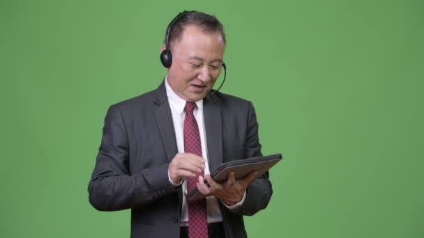 Mature Japanese businessman working as call center representative against green background — Stock Video