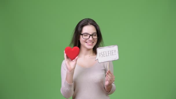 Joven feliz hermosa mujer sosteniendo corazón rojo y selfie papel signo — Vídeo de stock