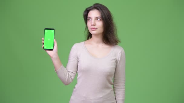 Молода красива жінка думає, показуючи телефон на зеленому тлі — стокове відео