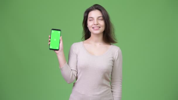 Joven feliz hermosa mujer pensando mientras muestra el teléfono contra el fondo verde — Vídeo de stock