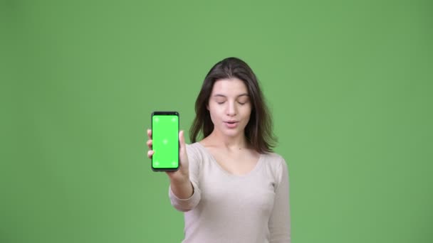 Jeune belle femme regardant choqué tout en montrant le téléphone sur fond vert — Video