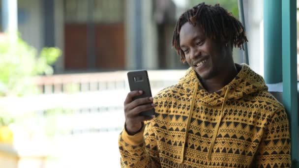 Joven hombre africano guapo feliz sonriendo mientras usa el teléfono en las calles al aire libre — Vídeo de stock