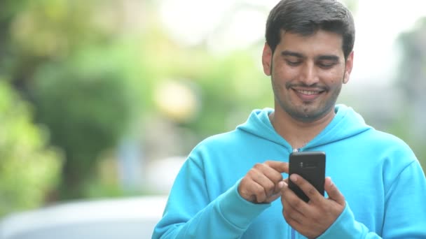 Mladý šťastný perský muž myšlení při používání telefonu venku — Stock video