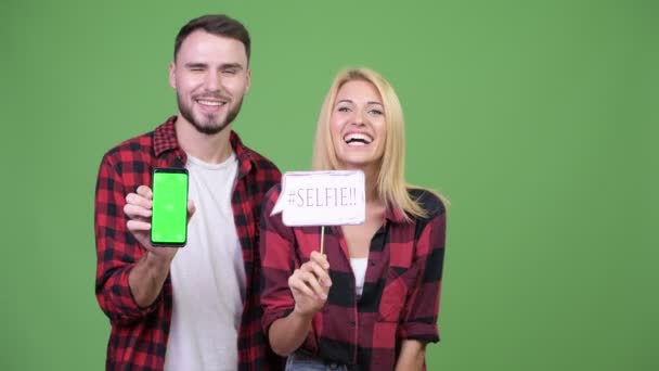 Pareja joven mostrando el teléfono mientras sostiene selfie papel signo juntos — Vídeo de stock