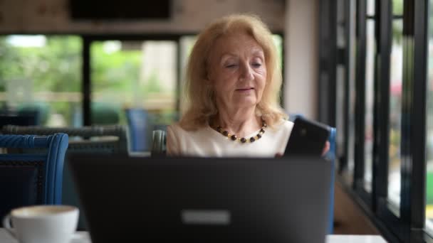 Mujer mayor utilizando ordenador portátil y hablando en el teléfono móvil — Vídeo de stock