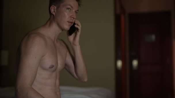 Мужчина без рубашки разговаривает по мобильному телефону ночью в постели в отеле — стоковое видео