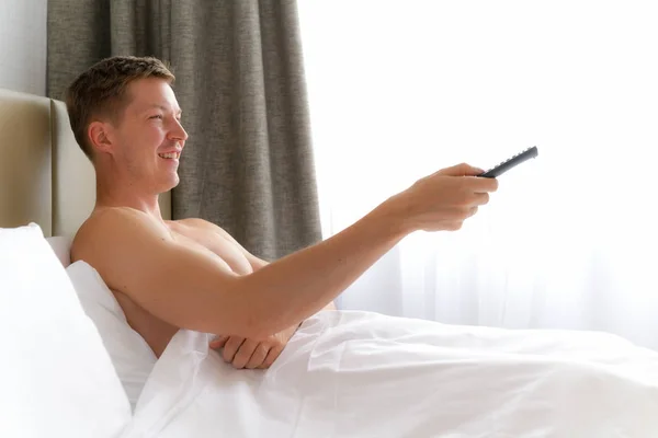 Homem na cama assistindo televisão e segurando TV remoto — Fotografia de Stock