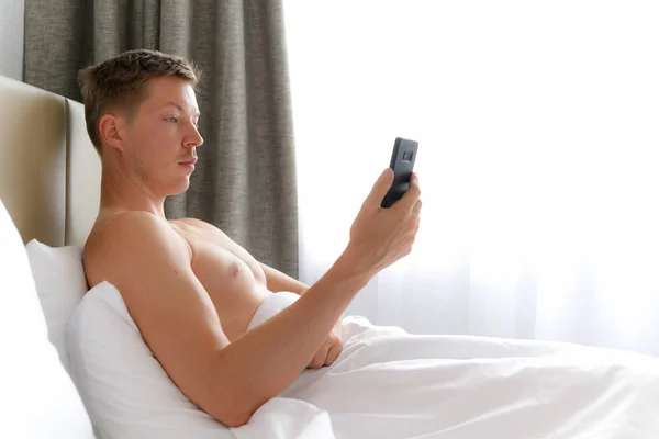 Giovane uomo senza maglietta utilizzando il telefono cellulare a letto — Foto Stock