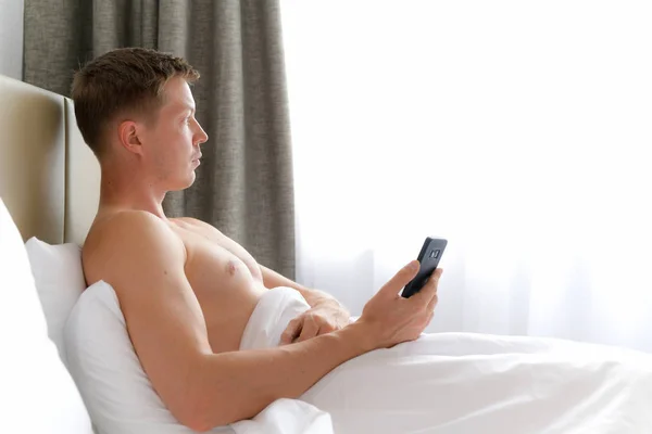 Uomo utilizzando il telefono cellulare a letto e guardando attraverso la finestra — Foto Stock