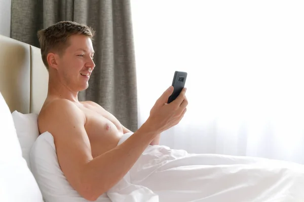 Jovem sem camisa homem usando celular na cama — Fotografia de Stock