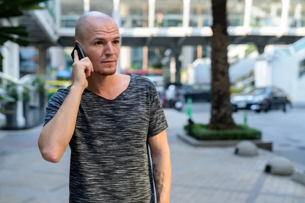 Young handsome bald man thinking while talking on mobile phone o