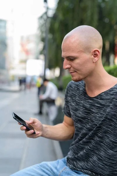 Profile view of young handsome bald man using mobile phone outsi — Stock Photo, Image