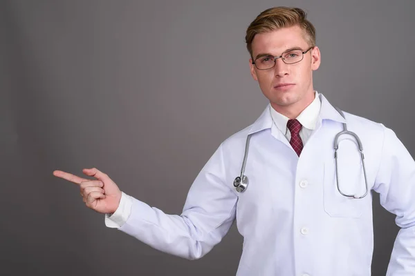 灰色の背景の眼鏡を身に着けている金髪のハンサムな青年医師のスタジオ撮影 — ストック写真