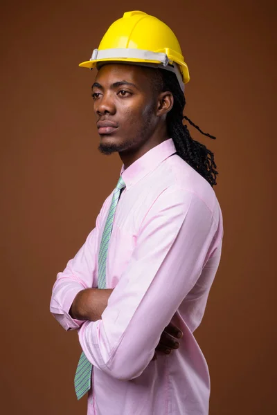 Joven hombre de negocios africano guapo con hardhat contra ba marrón —  Fotos de Stock