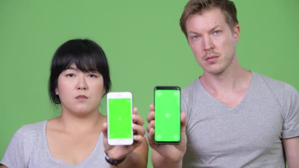 Pasangan multi-etnis muda yang bahagia menunjukkan dua ponsel bersama-sama — Stok Video