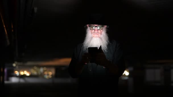 Зрілий бородатий чоловік виглядає підозріло під час використання телефону в темряві — стокове відео