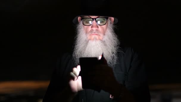 Зрелый бородатый мужчина выглядит подозрительно, когда пользуется телефоном в темноте — стоковое видео