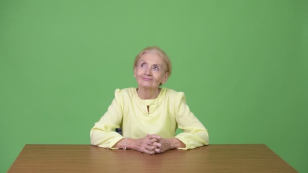 Красивая счастливая старшая деловая женщина улыбается, думая против деревянного стола — стоковое видео