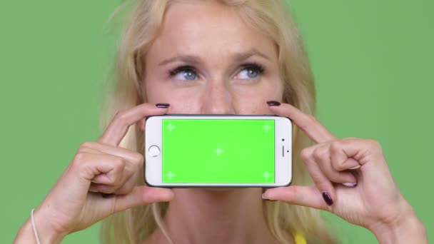 Jonge gelukkig mooie blonde vrouw die denken terwijl het tonen van de telefoon — Stockvideo