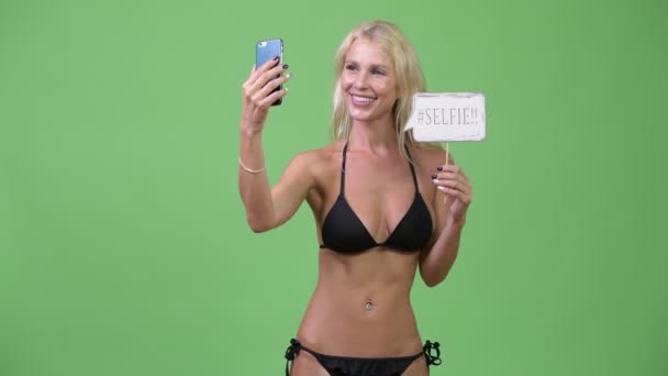 Lykkelig ung, blond kvinne i bikini mens hun tok selfie med papirskilt – stockvideo
