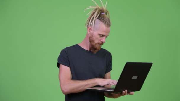 Щасливий красивий бородатий чоловік з дредлоками мислення під час використання ноутбука — стокове відео