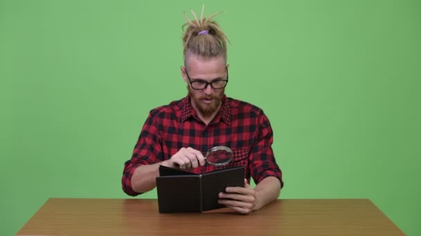 Guapo barbudo hombre hipster libro de lectura con lupa contra mesa de madera — Vídeo de stock