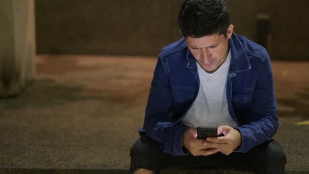 Hiszpanin człowieka przy użyciu telefonu siedząc na ulicy w mieście w nocy — Wideo stockowe