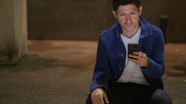 Hispanic man thinking and using phone while sitting on the street in the city at night — Stock Video