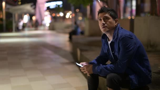 Spaanse man denken en met behulp van telefoon zittend op de straat van de stad bij nacht — Stockvideo