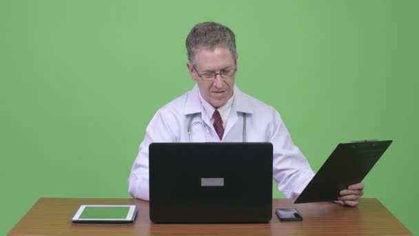 Porträtt av mogen man läkare multi-tasking arbete mot träbord — Stockvideo