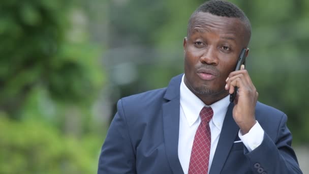 Молодой счастливый африканский бизнесмен думает, разговаривая по телефону на улице — стоковое видео