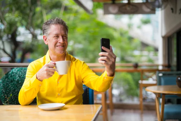 コーヒー ショップで携帯電話を使用して Selfie をしながら座っているハンサムな男を成熟します。 — ストック写真