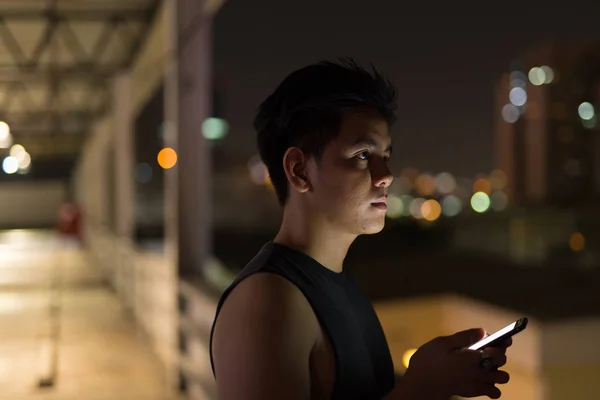 Όμορφος Ασίας νεαρός σκέψης κατά τη χρήση του τηλεφώνου κατά την άποψη της πόλης τη νύχτα — Φωτογραφία Αρχείου