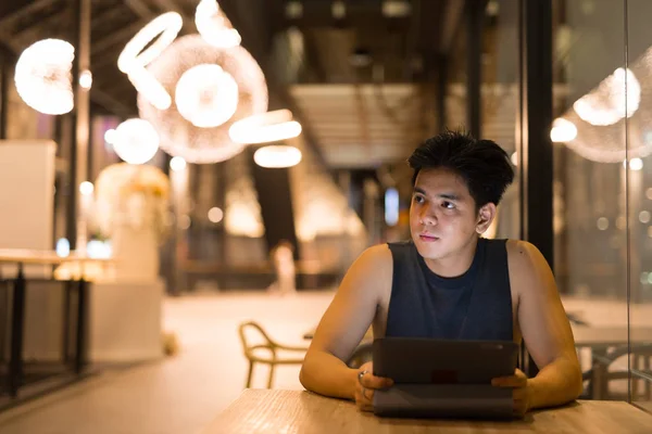 Joven guapo asiático hombre pensando mientras se utiliza la tableta digital en el restaurante por la noche — Foto de Stock