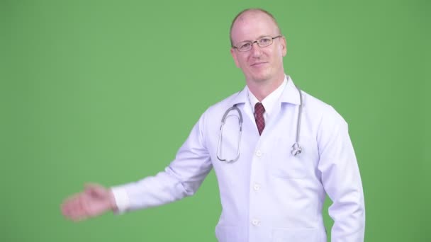 Feliz homem careca maduro médico mostrando algo contra fundo verde — Vídeo de Stock