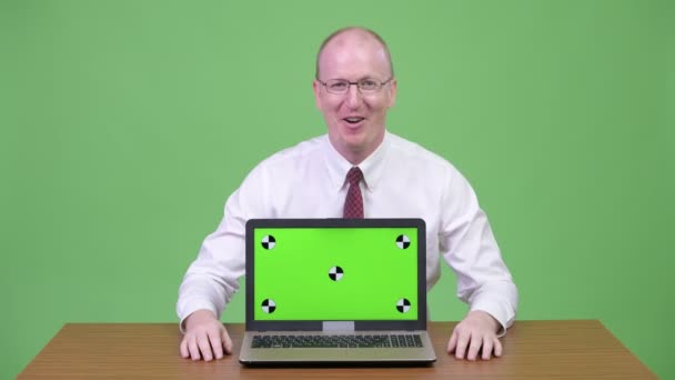Happy mature bald businessman promoting while showing laptop against wooden table — Stock Video