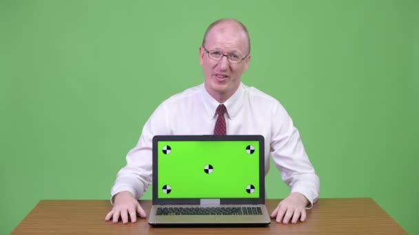 Оброблений зрілий лисий бізнесмен показує погані новини на ноутбуці проти дерев'яного столу — стокове відео