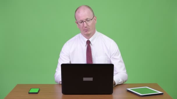 Зрелый лысый бизнесмен, использующий ноутбук и телефон на работе против деревянного стола — стоковое видео