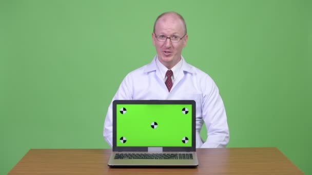 Doktor šťastný starší plešatý muž mluví zároveň ukazuje notebook proti dřevěný stůl — Stock video
