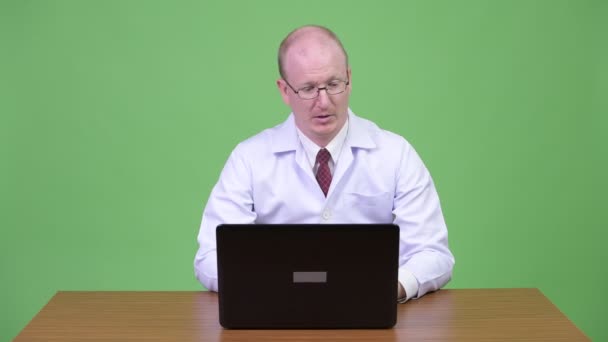 Stressed mature bald man doctor arguing to the laptop against wooden table — Stock Video