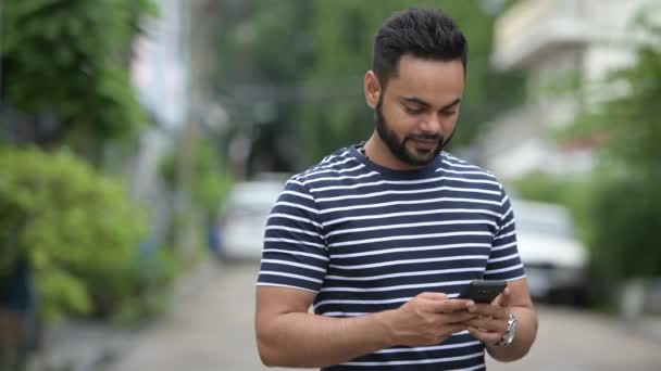 Jeune homme indien barbu heureux en utilisant le téléphone en plein air — Video