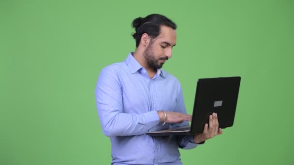 Молодой счастливый бородатый индийский бизнесмен думает во время использования ноутбука — стоковое видео