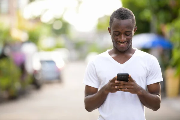 Jovem homem africano feliz sorrindo ao usar o telefone nas ruas ao ar livre — Fotografia de Stock