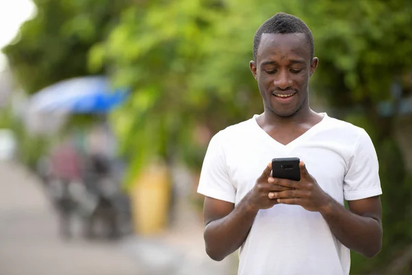 Jovem homem africano feliz sorrindo ao usar o telefone nas ruas ao ar livre — Fotografia de Stock